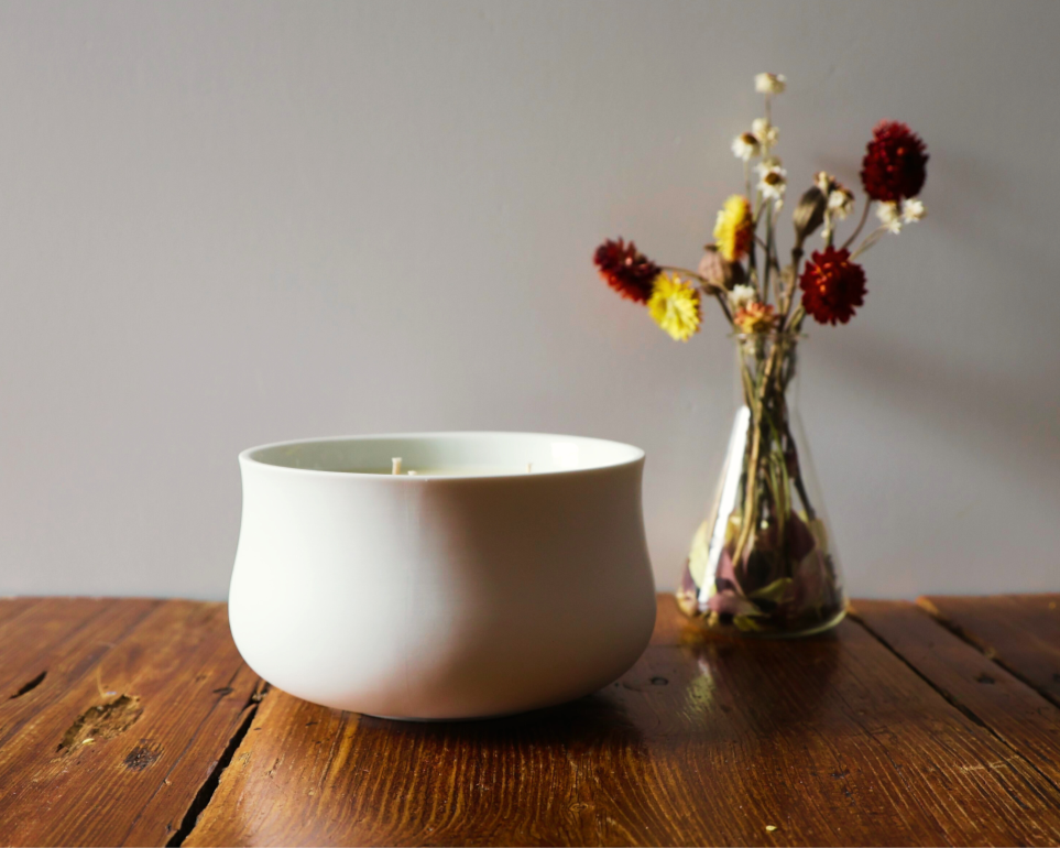 A minimalist candle in a white bowl next to a vase of dried flowers on a wooden table.