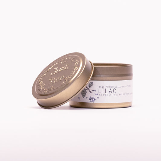 Just Bee Lilac travel candle in a gold tin with the embossed Just Bee lid propped against it.