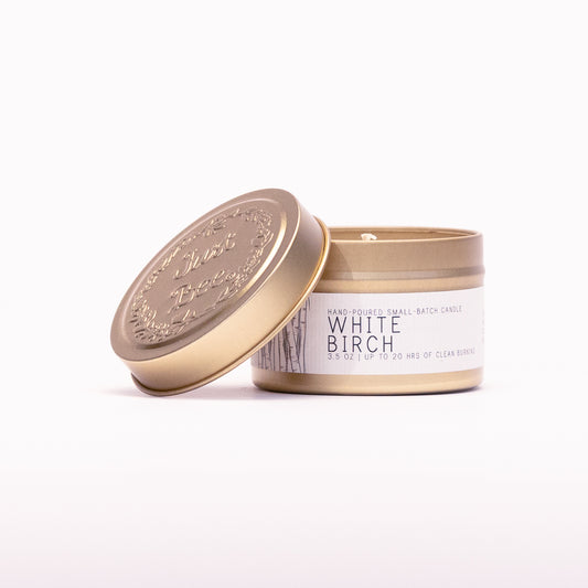 Just Bee White Birch travel candle in a gold tin with the embossed Just Bee lid propped against it.