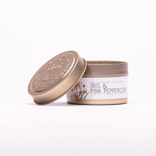 Just Bee Iris & Pink Peppercorn travel candle in a gold tin with the embossed Just Bee lid propped beside it.
