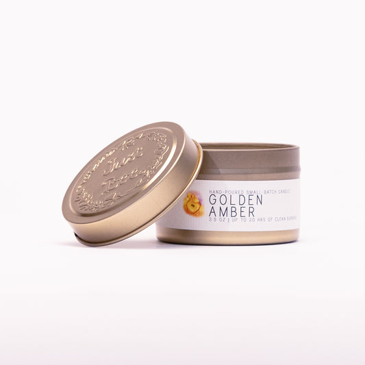 Just Bee Golden Amber travel candle in a gold tin with the embossed Just Bee lid propped against it.
