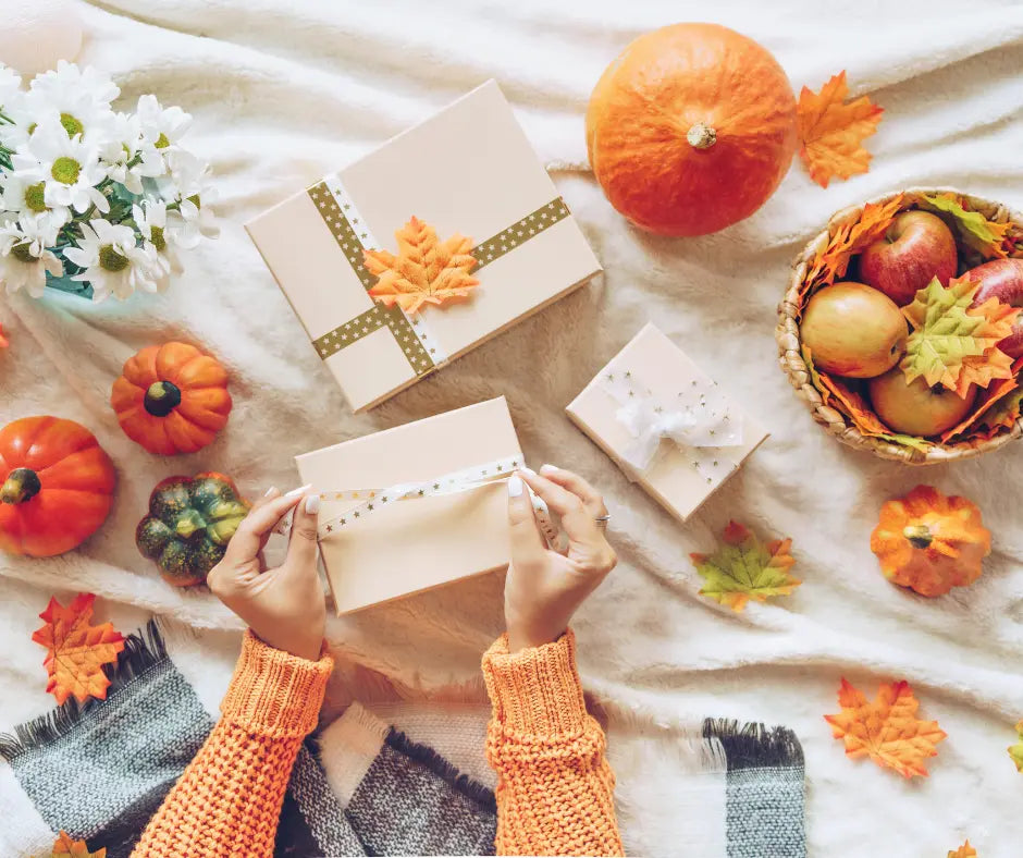 Hands wrapping gift boxes with autumn leaves, pumpkins, apples, and white flowers styled around them for teacher gifts