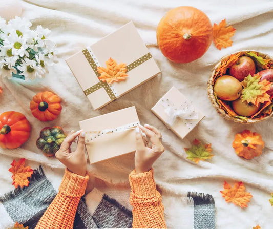 Hands wrapping gift boxes with autumn leaves, pumpkins, apples, and white flowers styled around them for teacher gifts