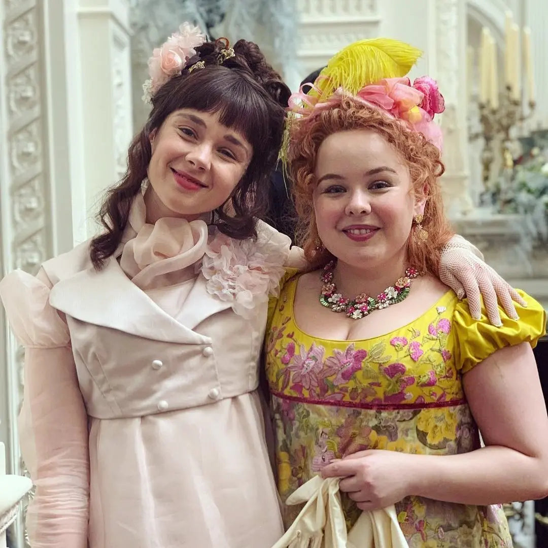 Two women in elaborate Regency-era costumes with floral headpieces smiling together, styled after the show Bridgerton