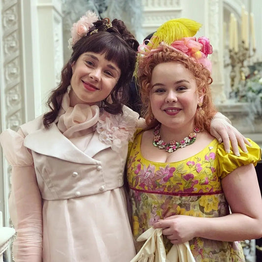 Two women in elaborate Regency-era costumes with floral headpieces smiling together, styled after the show Bridgerton
