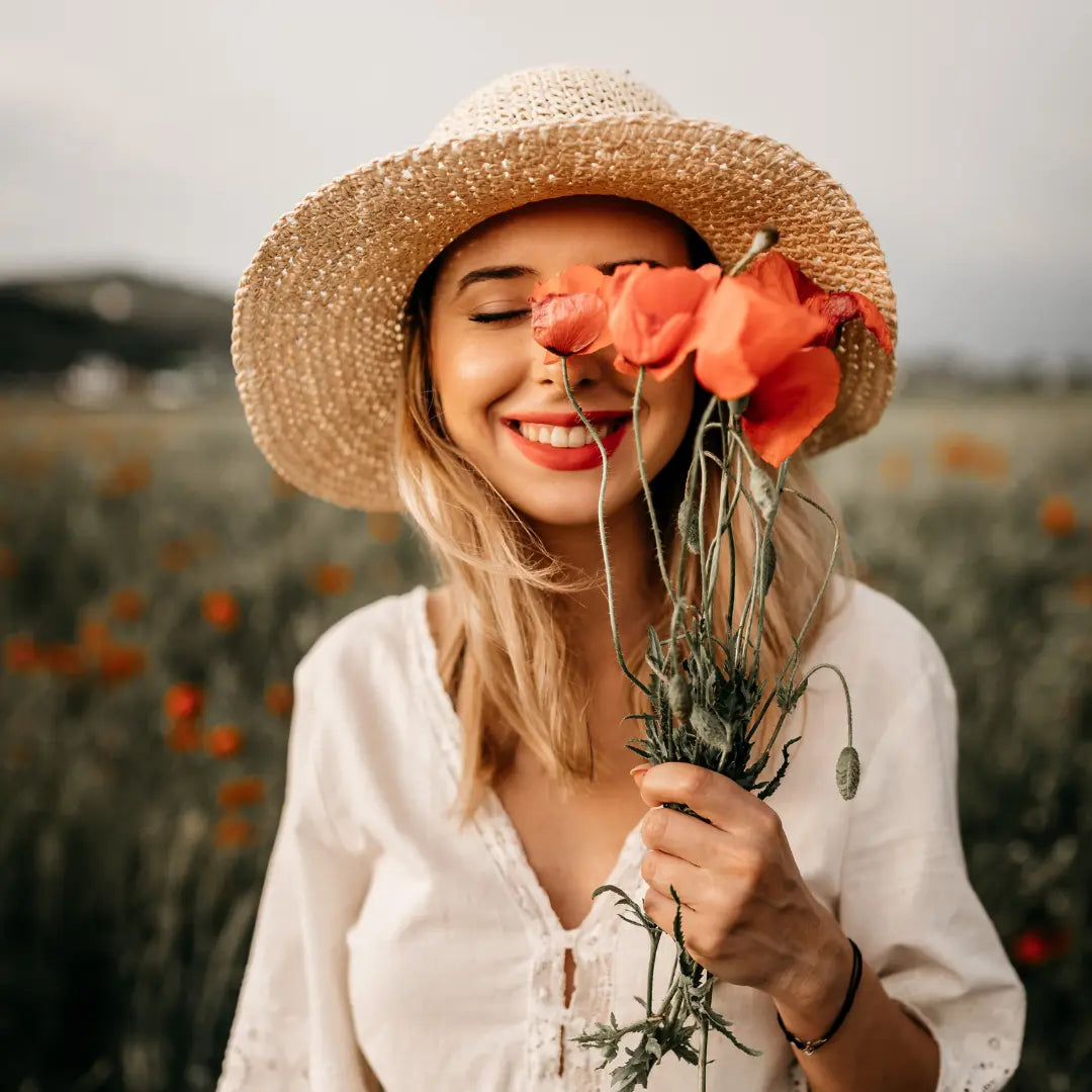 A smiling woman in a straw hat holding orange poppies up to her face in a summer field
