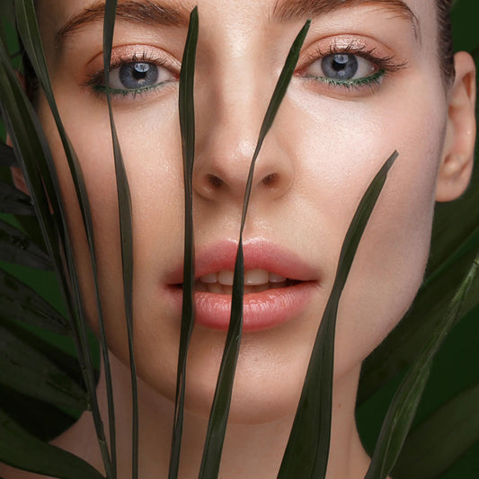 Close-up of a woman's face with green eyeliner and glossy lips, peeking through green palm leaves