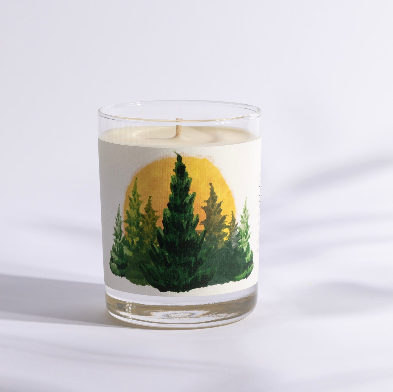 A candle in a glass container featuring a design of trees and a yellow sun.