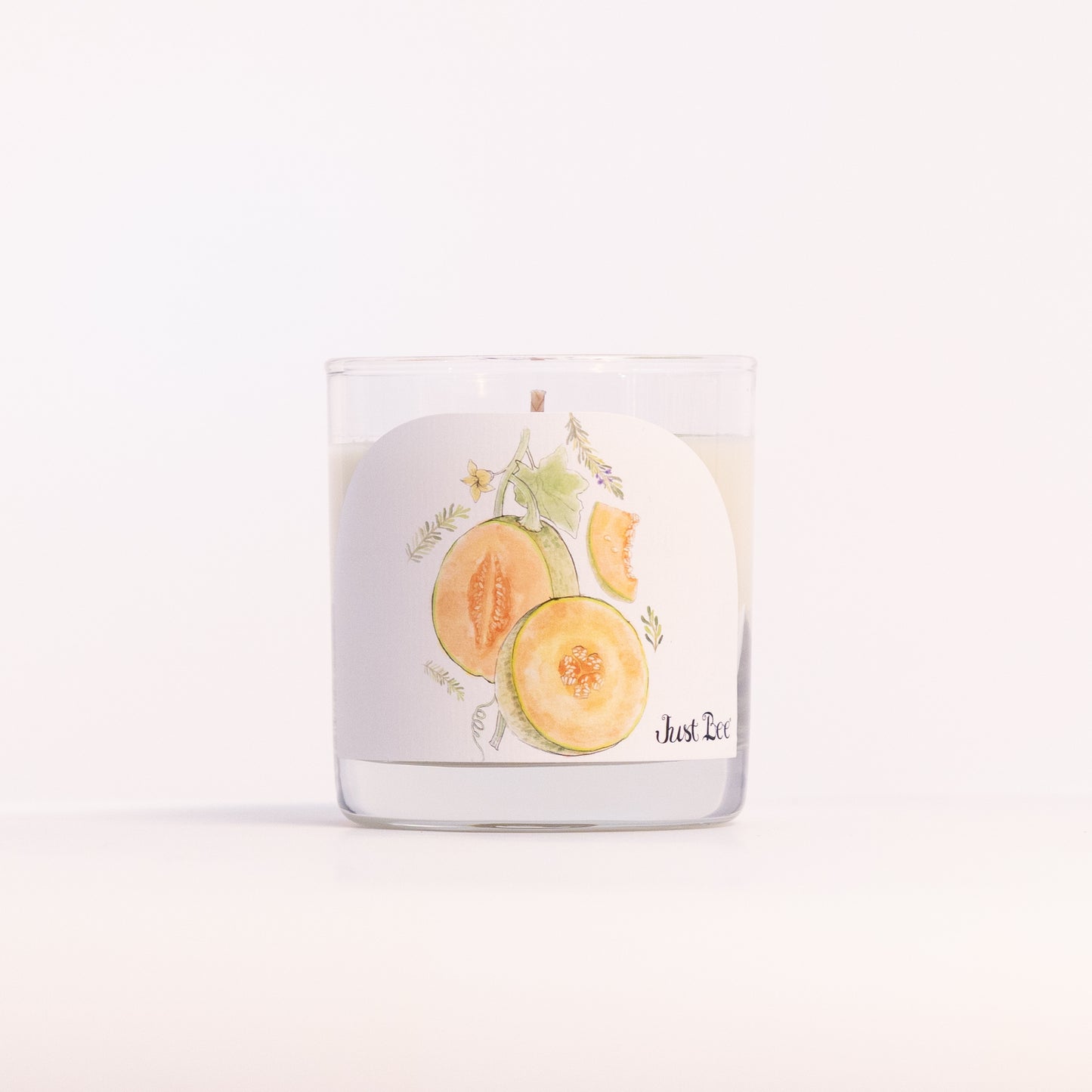 Just Bee Persian Melon & Rosemary candle in a clear glass jar, melon label facing forward, closer view.
