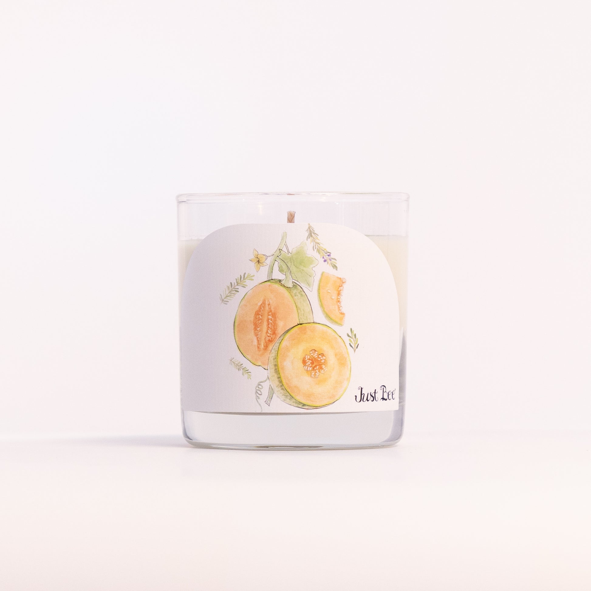 Just Bee Persian Melon & Rosemary candle in a clear glass jar, melon label facing forward, closer view.