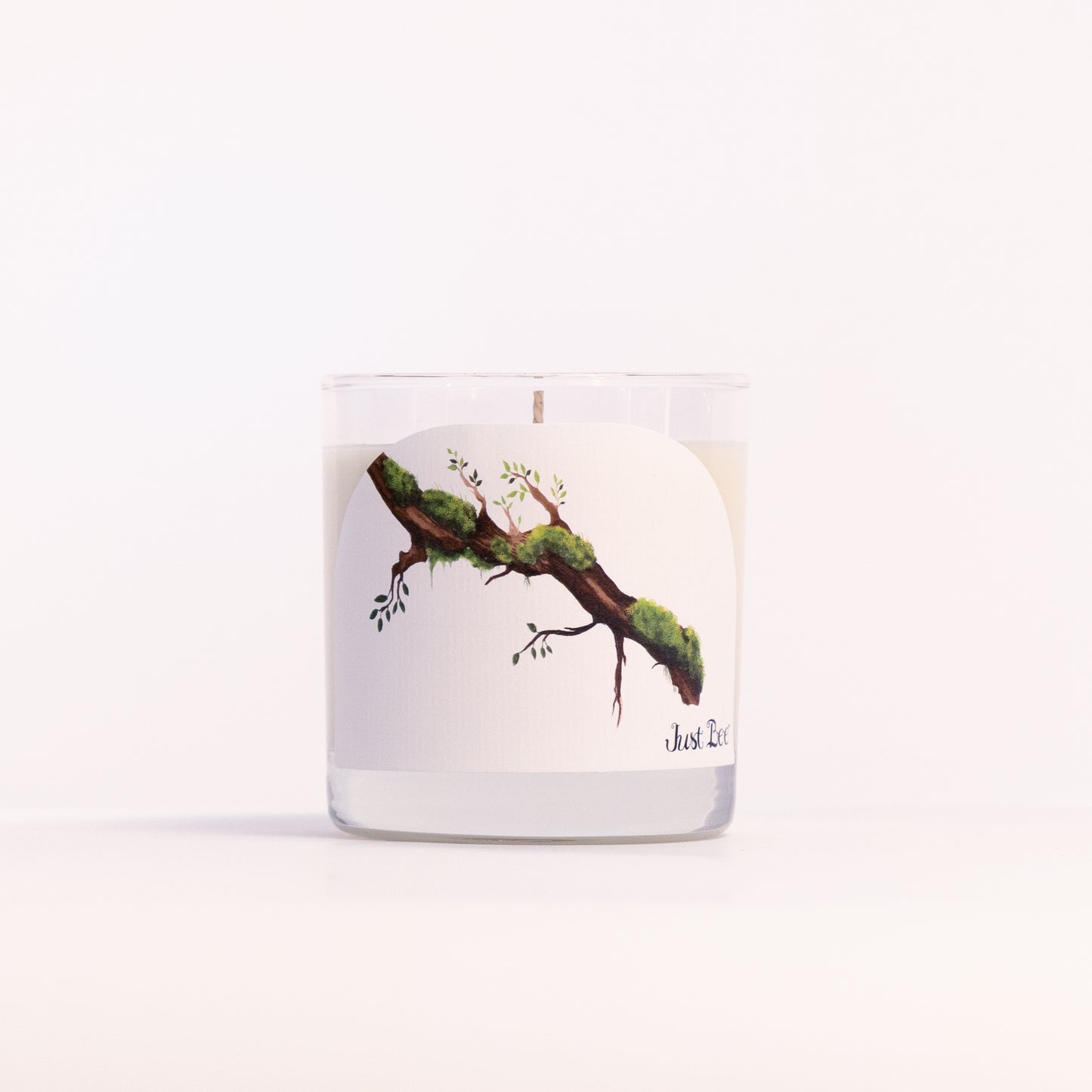 Just Bee Guaiac Wood & Moss candle in a clear glass jar, mossy branch label facing forward, closer view.