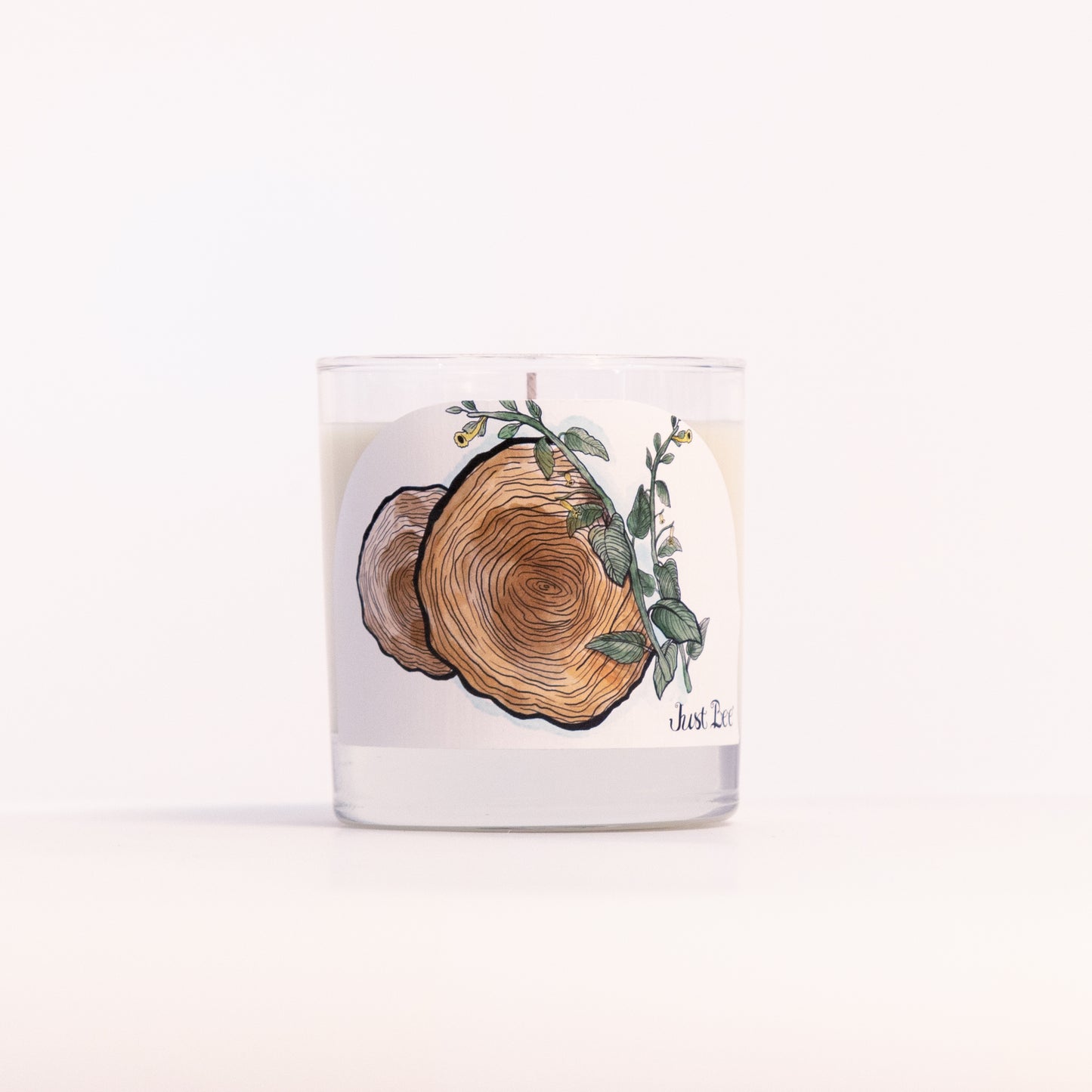 Just Bee Teakwood Tobacco candle in a clear glass jar, wood slice label facing forward, closer view.