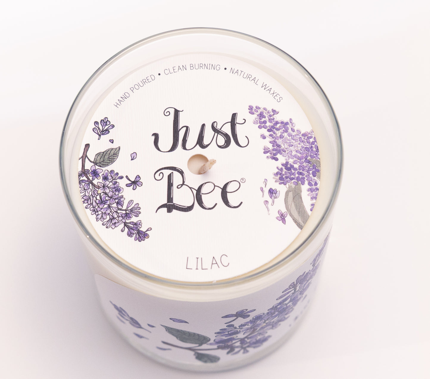 Just Bee Lilac candle from above, round lid label with purple lilac blossoms and a centered wick.