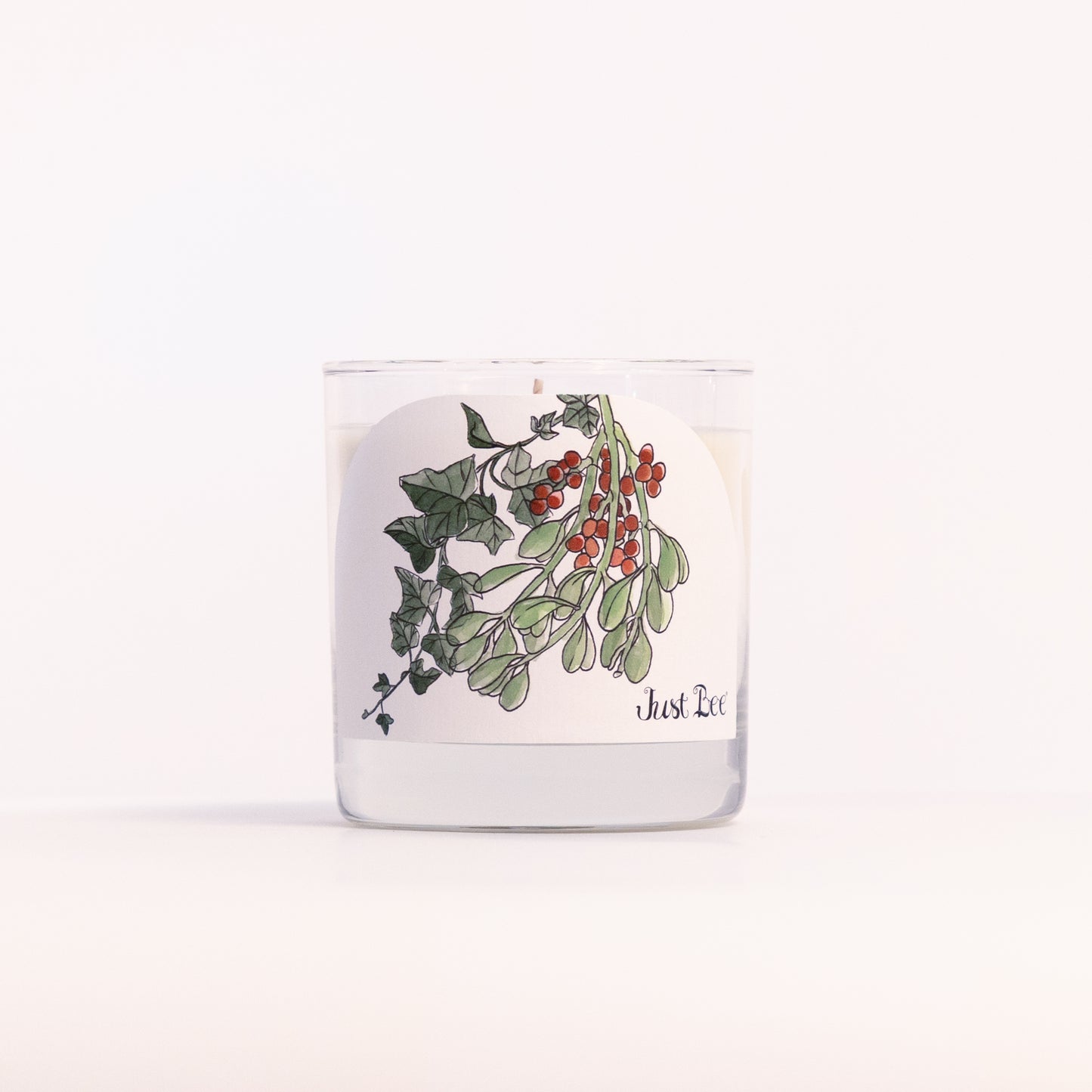 Just Bee Holly & Ivy candle in a clear glass jar, holly and berry label facing forward, closer view.