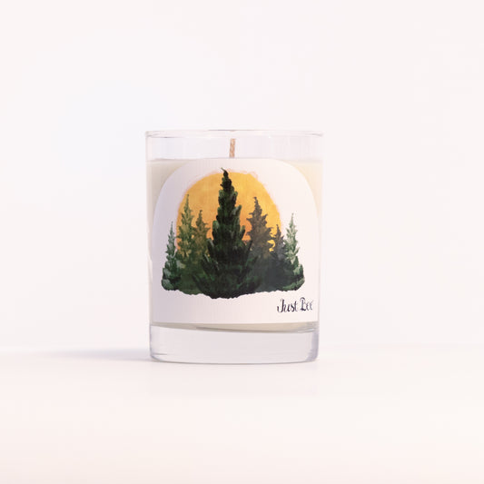 Just Bee Hinoki Sanctuary candle in a clear glass jar illustrated with evergreen pines and a golden sun.