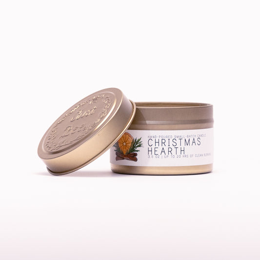 Just Bee Christmas Hearth travel candle in a gold tin with the embossed Just Bee lid propped against it.