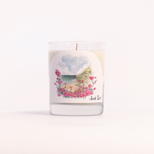 Just Bee Rockrose de la Mer candle in a clear glass jar illustrated with pink rockrose over a seascape.