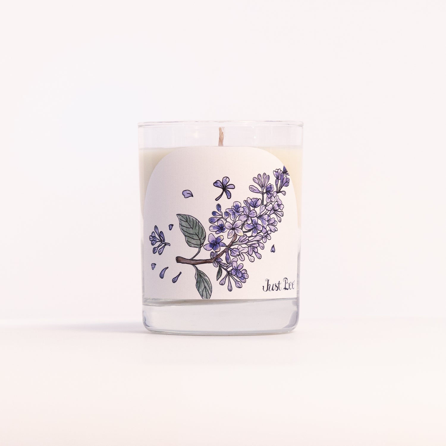Just Bee Lilac candle in a clear glass jar illustrated with a cluster of purple lilac blossoms.