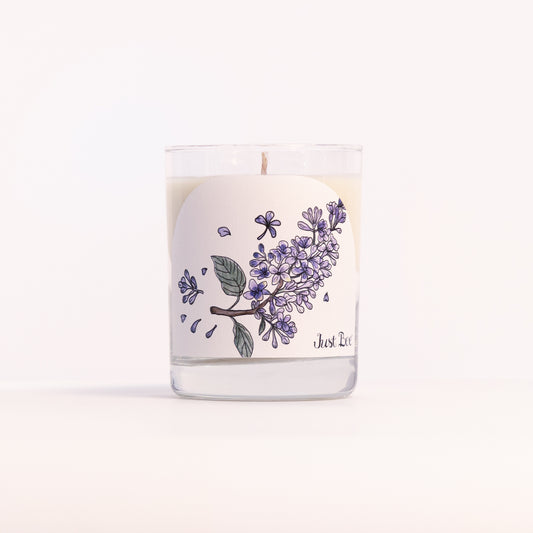 Just Bee Lilac candle in a clear glass jar illustrated with a cluster of purple lilac blossoms.