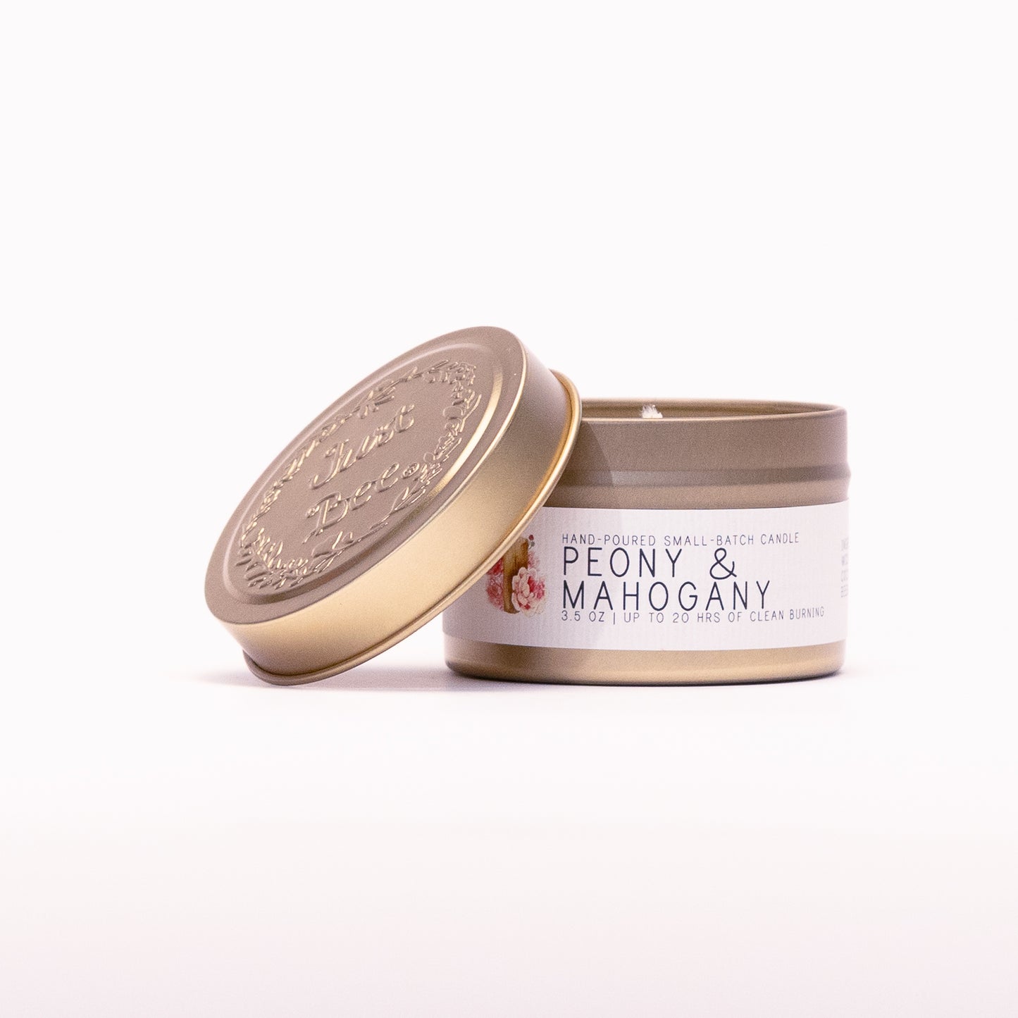 Just Bee Peony & Mahogany travel candle in a gold tin with the embossed Just Bee lid propped against it.