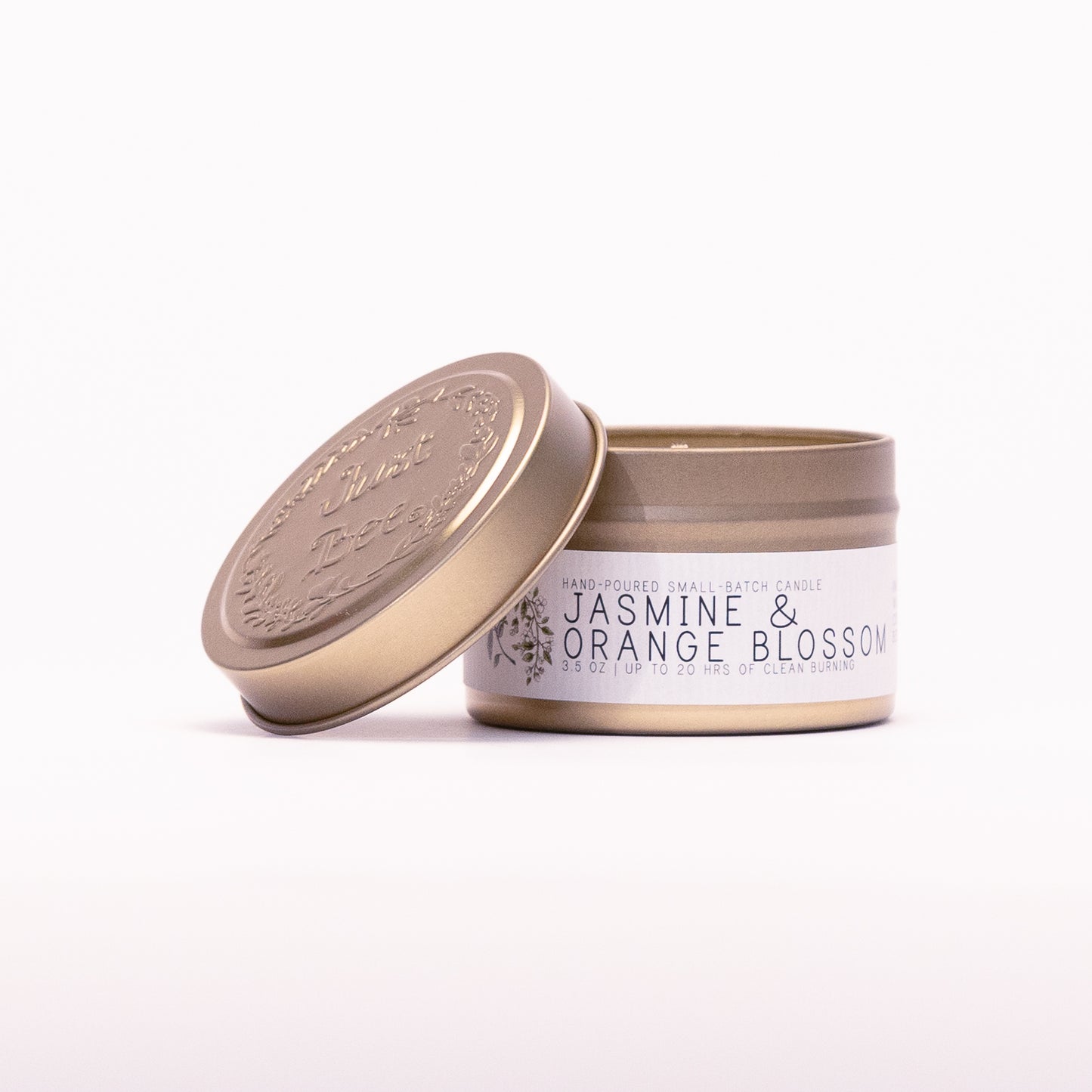 Just Bee Jasmine & Orange Blossom travel candle in a gold tin with the embossed Just Bee lid propped beside it.