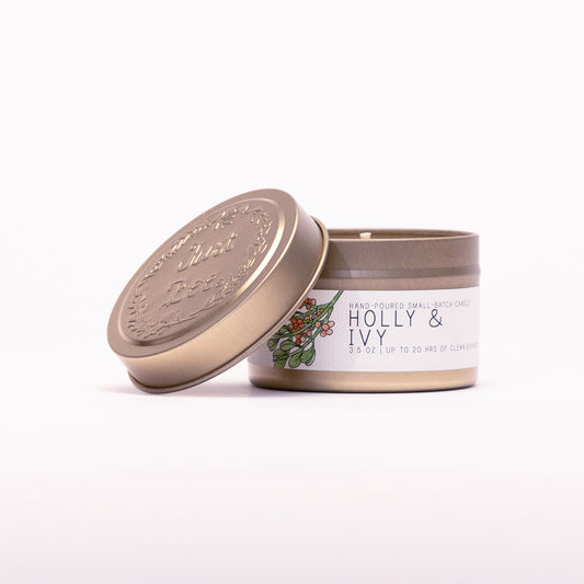 Just Bee Holly & Ivy travel candle in a gold tin with the embossed Just Bee lid propped against it.