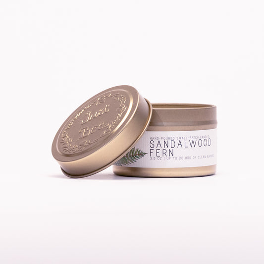 Just Bee Sandalwood Fern travel candle in a gold tin with the embossed Just Bee lid propped against it.