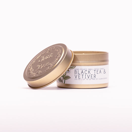 Just Bee Black Tea & Vetiver travel candle in a gold tin with the embossed Just Bee lid propped against it.