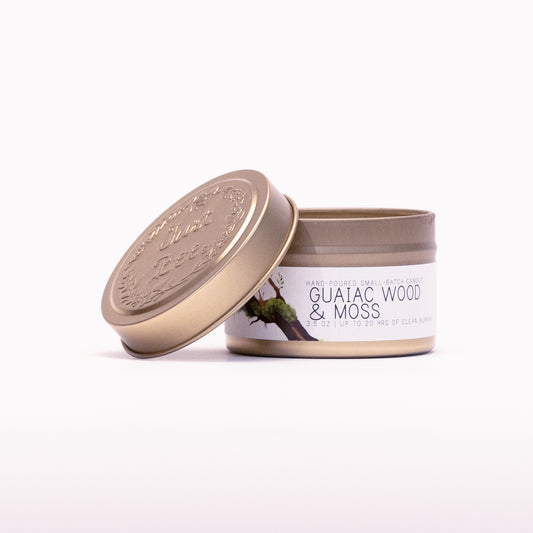 Just Bee Guaiac Wood & Moss travel candle in a gold tin with the embossed Just Bee lid propped against it.