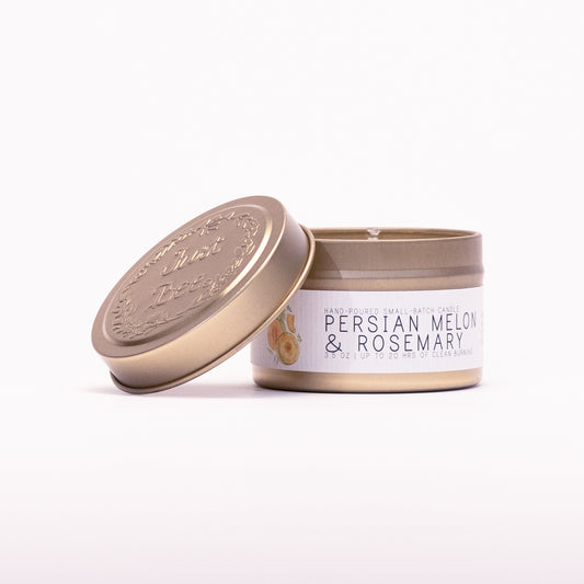 Just Bee Persian Melon & Rosemary travel candle in a gold tin with the embossed Just Bee lid propped beside it.
